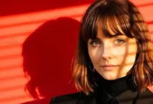 Woman with shoulder-length brown hair and bangs stands against a red wall with striped shadows across her face; wearing a black turtleneck and blazer.