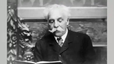 Elderly man with a white mustache in a suit reading an open book (black-and-white photo).