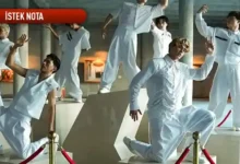 Dancers in white jumpsuits perform a contemporary routine around a geometric pedestal in a modern indoor space, with a red banner labeled 'İSTEK NOTA' visible above them.
